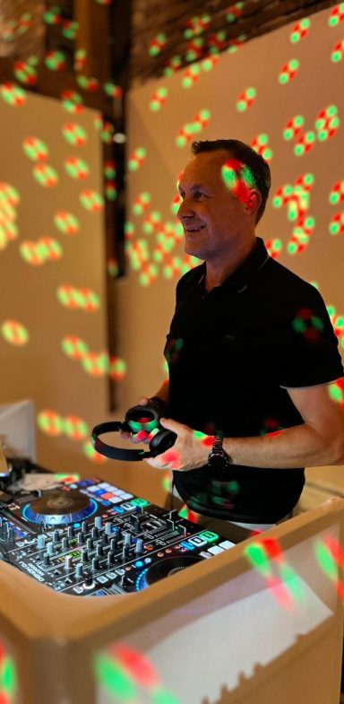 DJ steht an einem Mixer, umgeben von farbigen Lichtpunkten auf einer Wand.